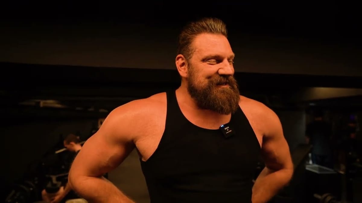 Olivier Richters in a black tank top, hands on hips, in his gym in warm low light