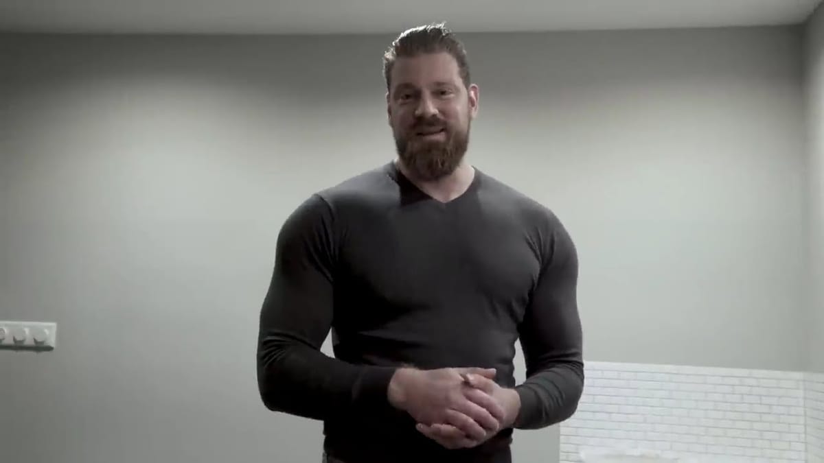 Olivier Richters standing, hands clasped, in a charcoal long-sleeve top in a plain grey room